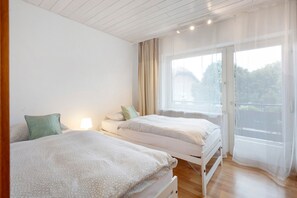 A bedroom with two beds, a window with sheer curtains, and a lamp on the nightstand.