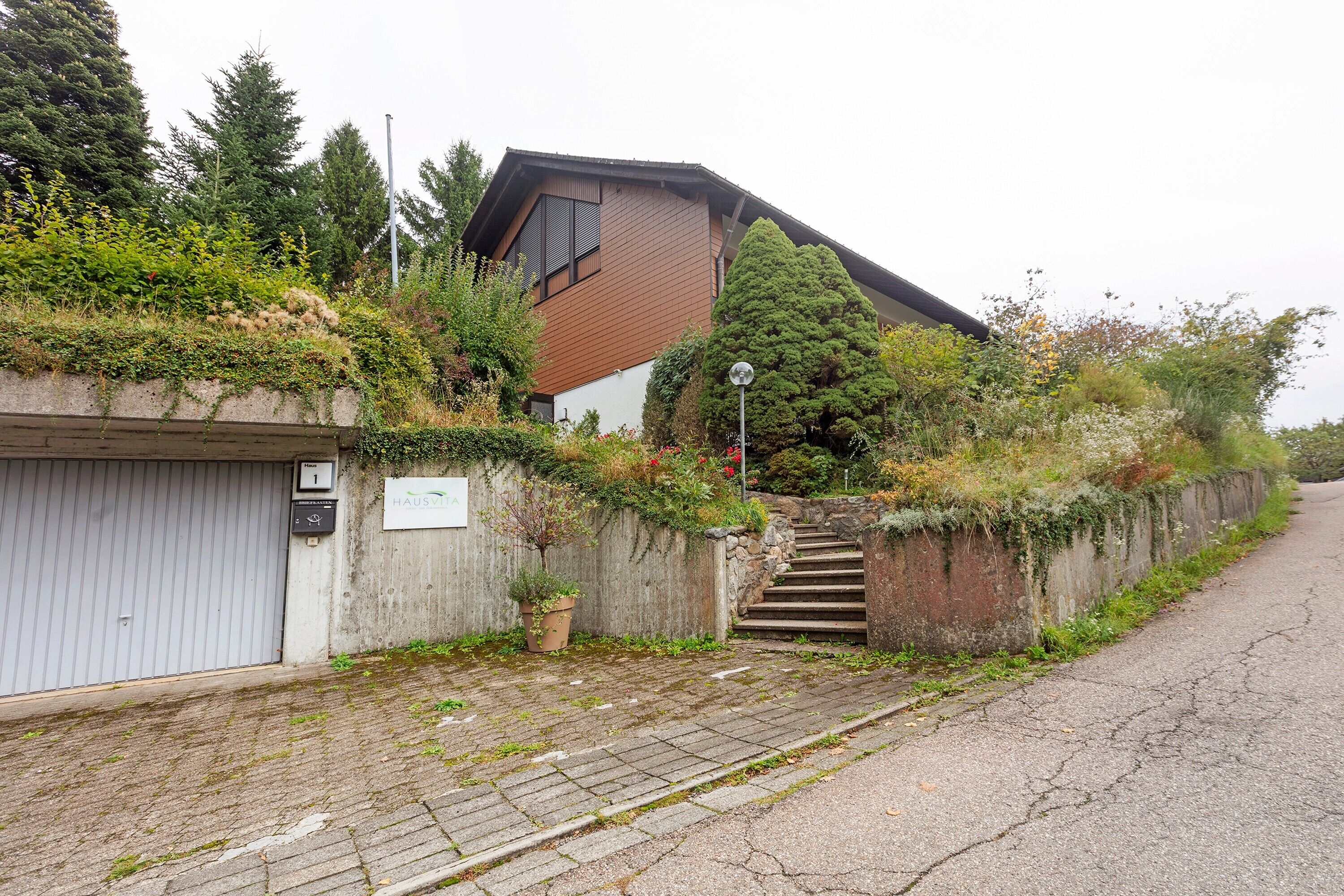 A residential building with a sloped roof, a staircase leading to a garage, and a paved driveway.