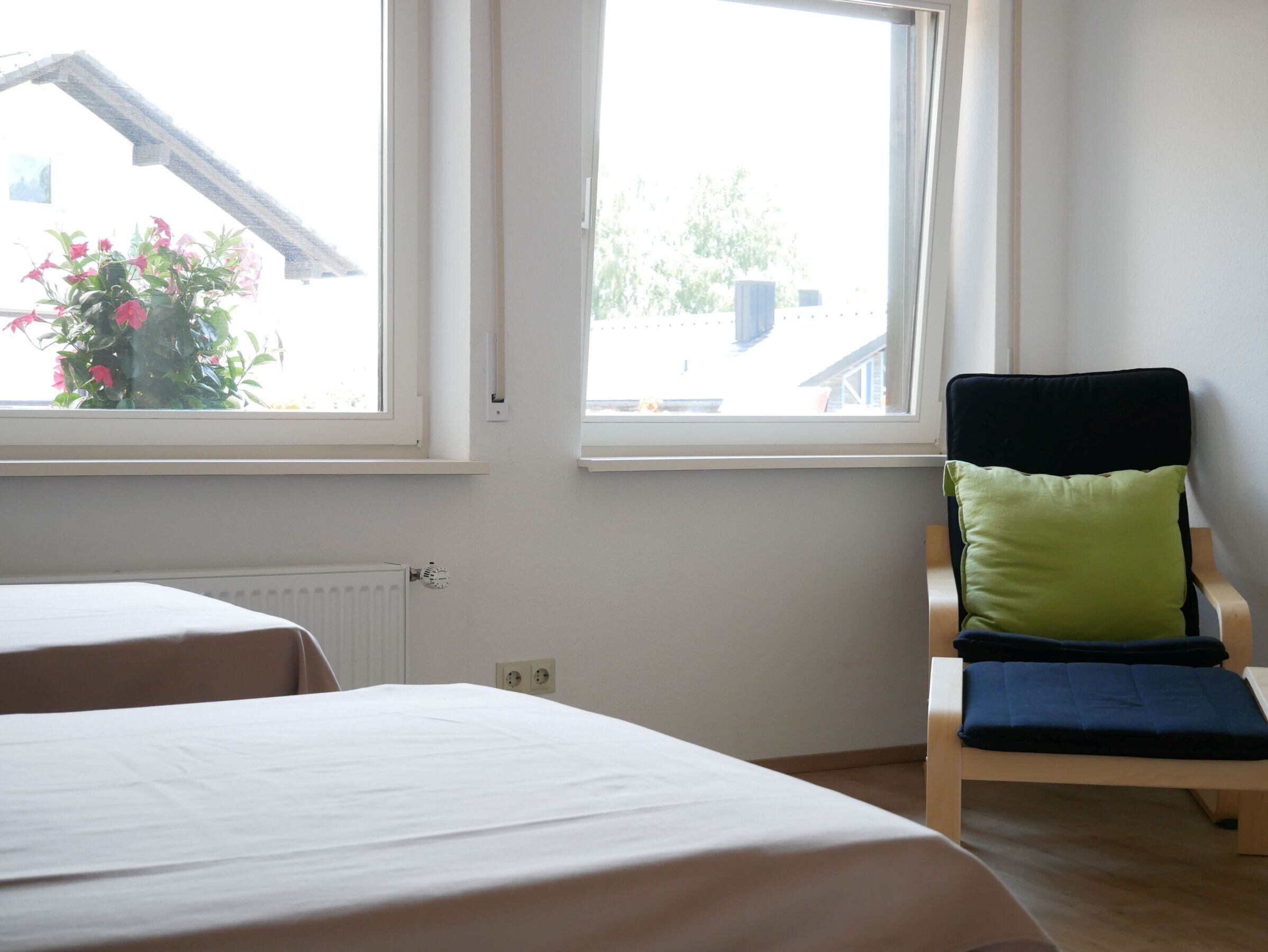 Een kamer met twee bedden, een fauteuil met kussens, en grote ramen met uitzicht op gebouwen en groen.
