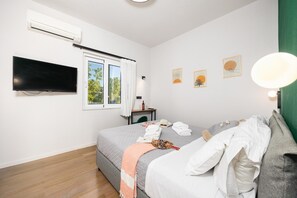 Un dormitorio con cama, televisión, ventana con cortinas, una mesita con una botella y decoración en la pared.