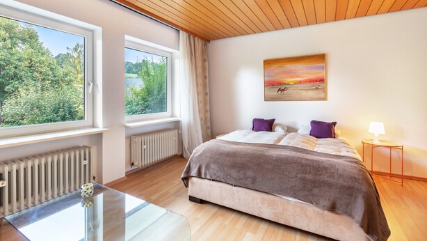 A bedroom with a large bed, a glass table, a window with a view of greenery, and a painting on the wall.