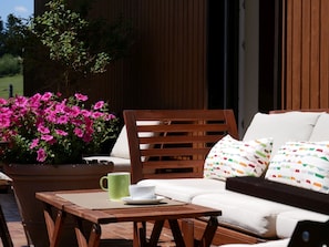 Un espace de détente extérieur en bois, comprenant une table, une chaise et un canapé agrémenté de coussins à motifs.