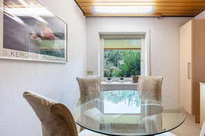Un coin repas avec une table et des chaises en verre, une fenêtre ornée de plantes et une photo encadrée accrochée au mur.