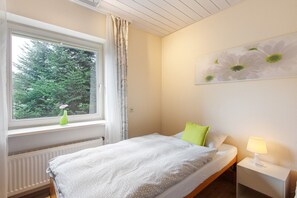 A bedroom with a bed, a nightstand with a lamp, a window with a view of trees, and a wall with a flower painting.