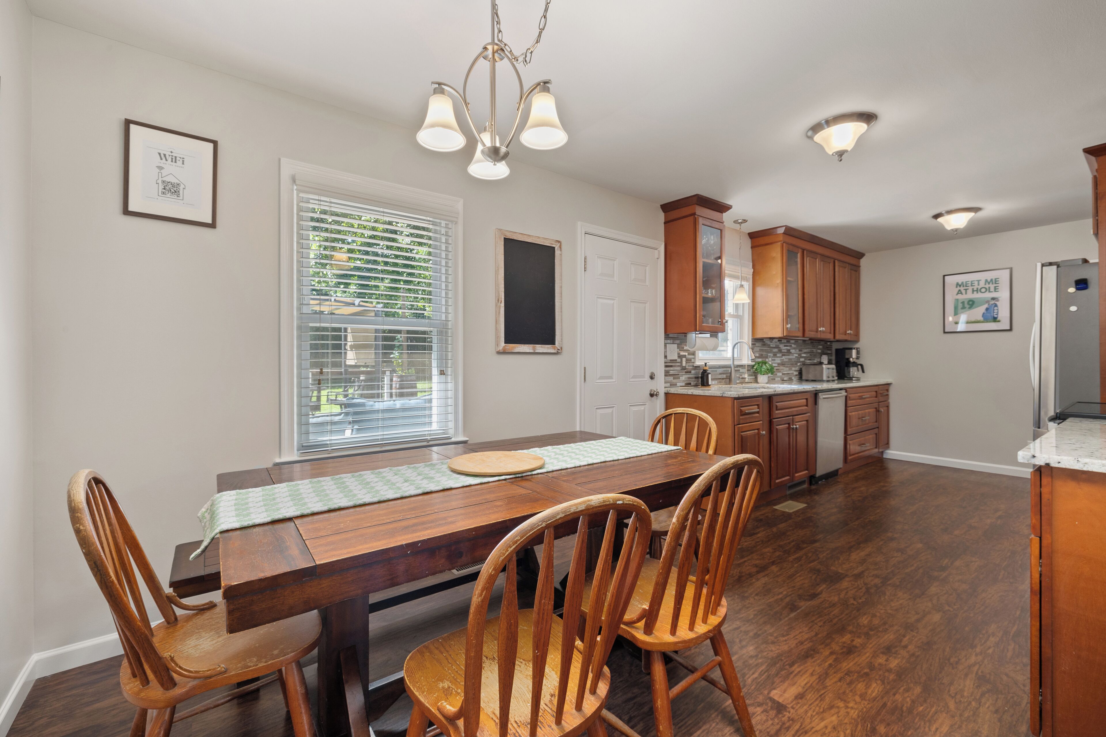 A dining area with a wooden table and chairs, a chandelier, and a kitchen in the background.