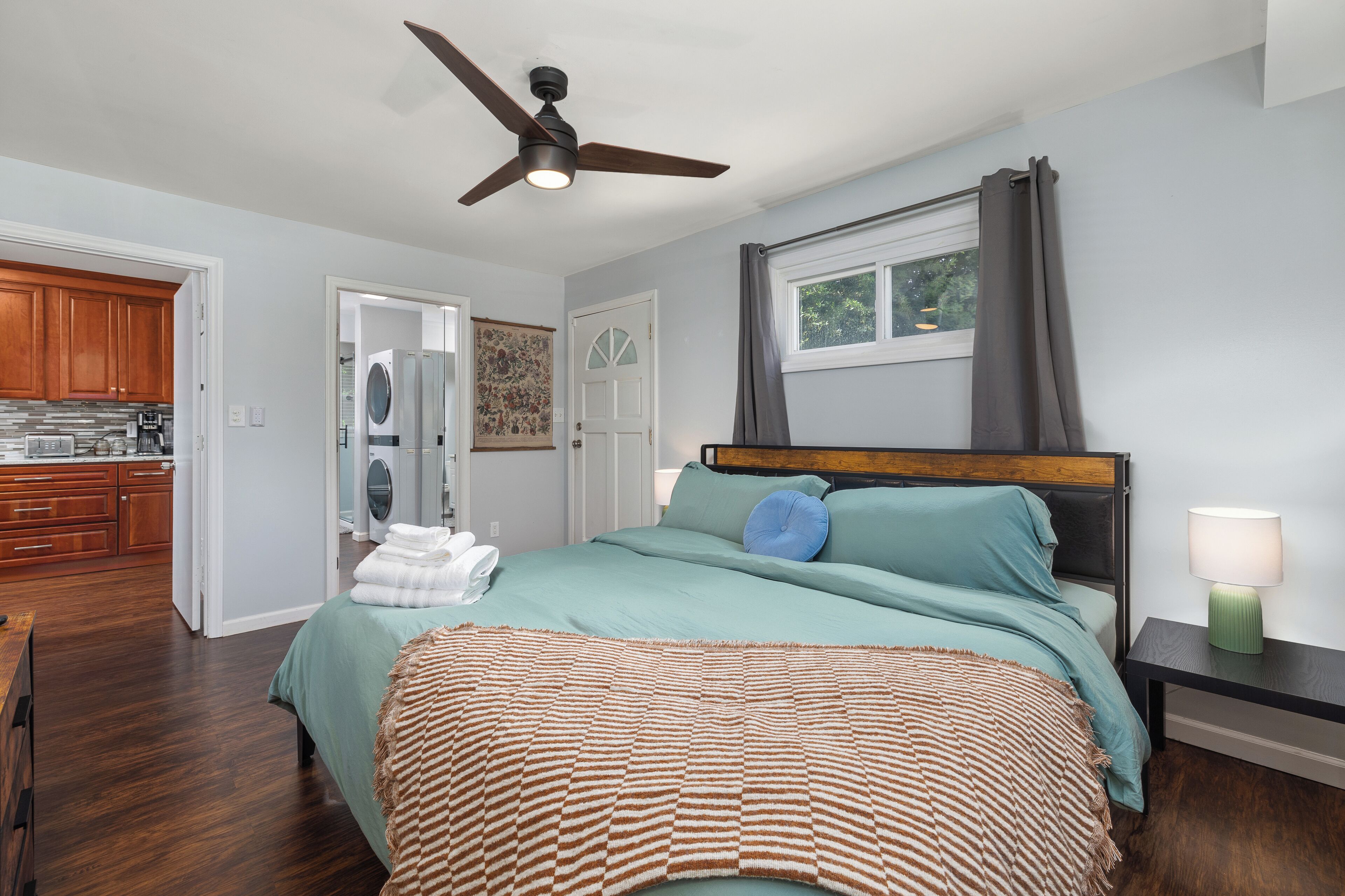 A bedroom with a bed, a nightstand, a ceiling fan, a window with curtains, and a view of the kitchen.