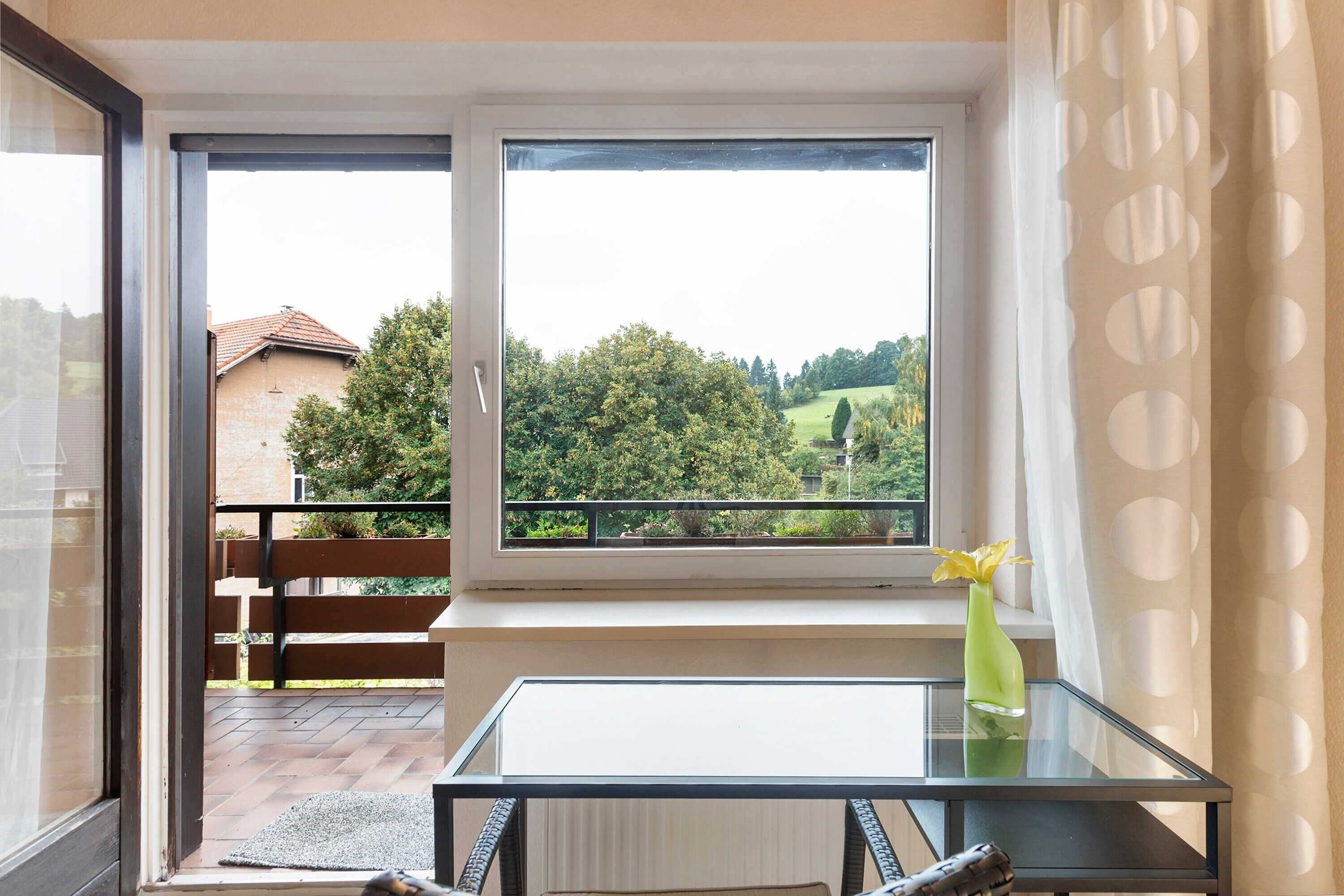 Ein Zimmer mit Glastisch, Bank, Fenster und Blick ins Grüne sowie einer Vase mit einer Blume.