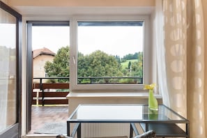 A room with a glass table, a bench, a window with a view of greenery, and a vase with a flower.