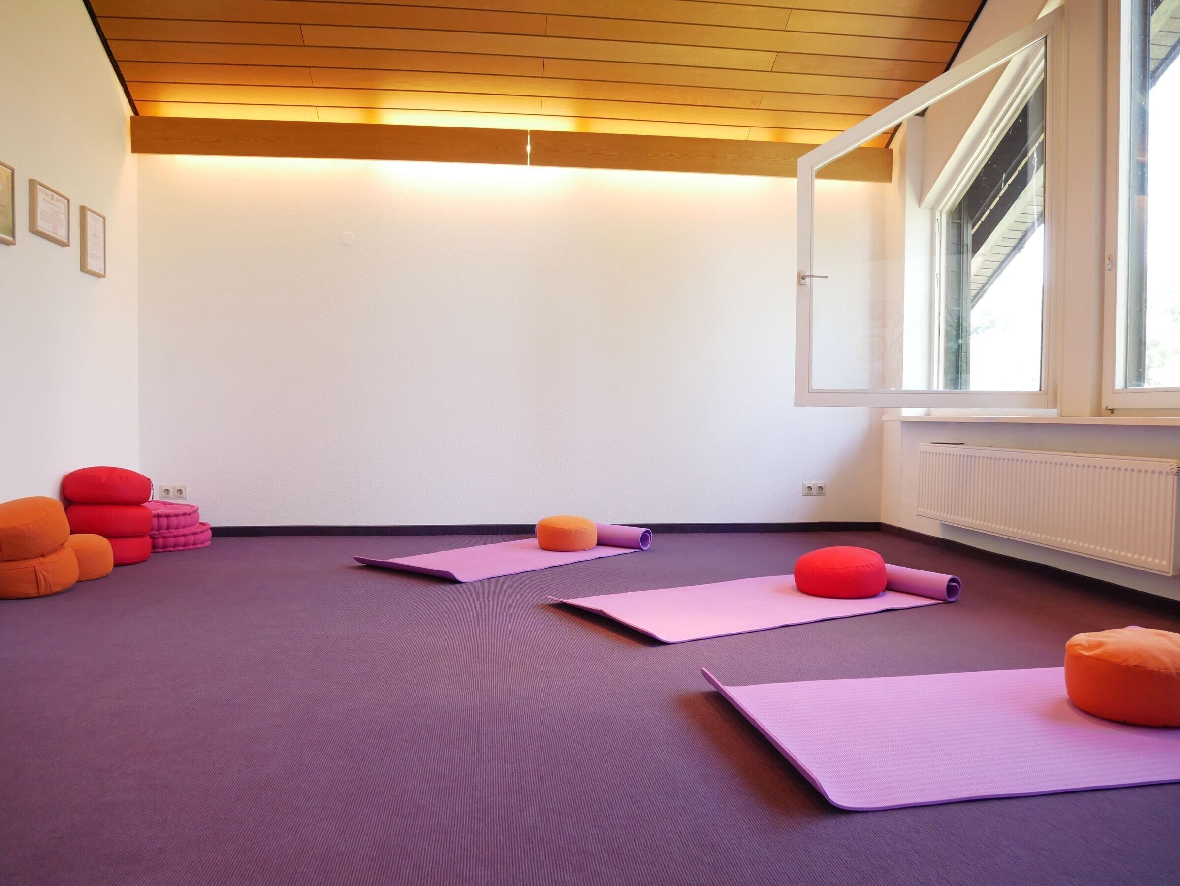Ein Yoga-Raum mit violetten Matten, orange- und roten Kissen und einem Fenster mit Blick.