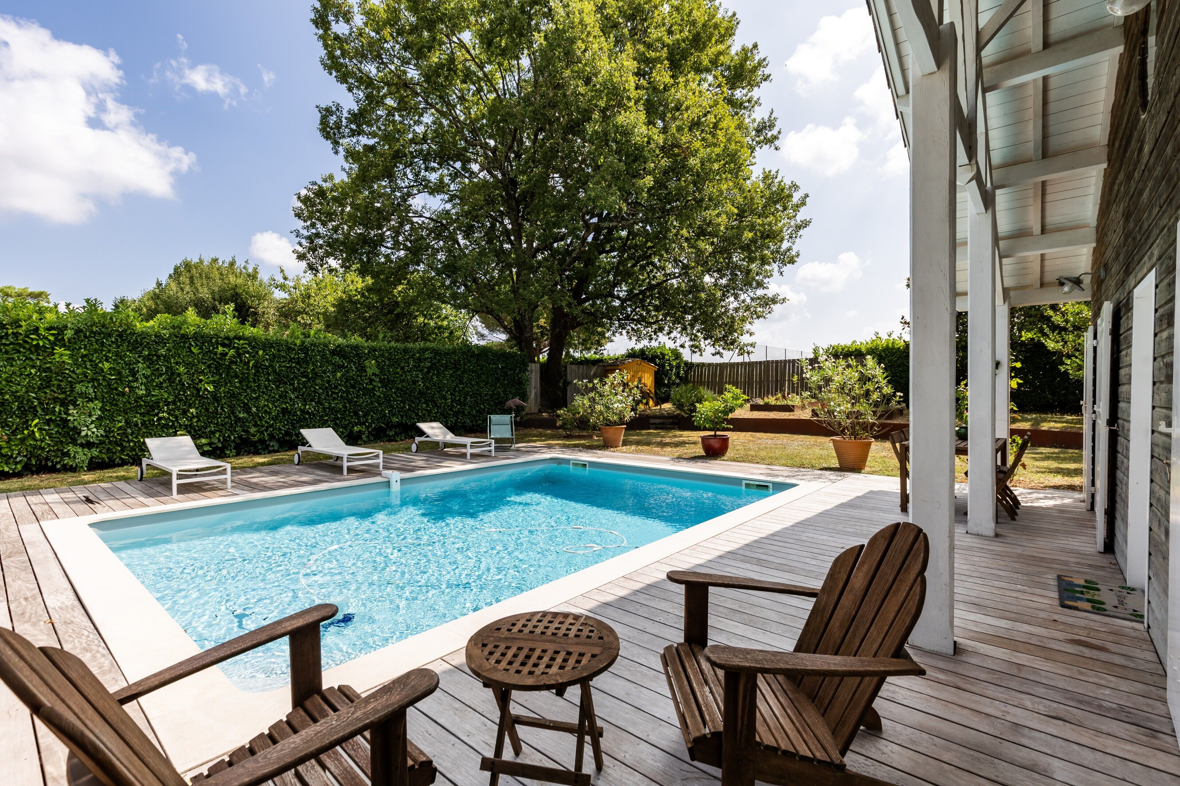 A rectangular swimming pool with a wooden deck, lounge chairs, and a covered area with wooden furniture.