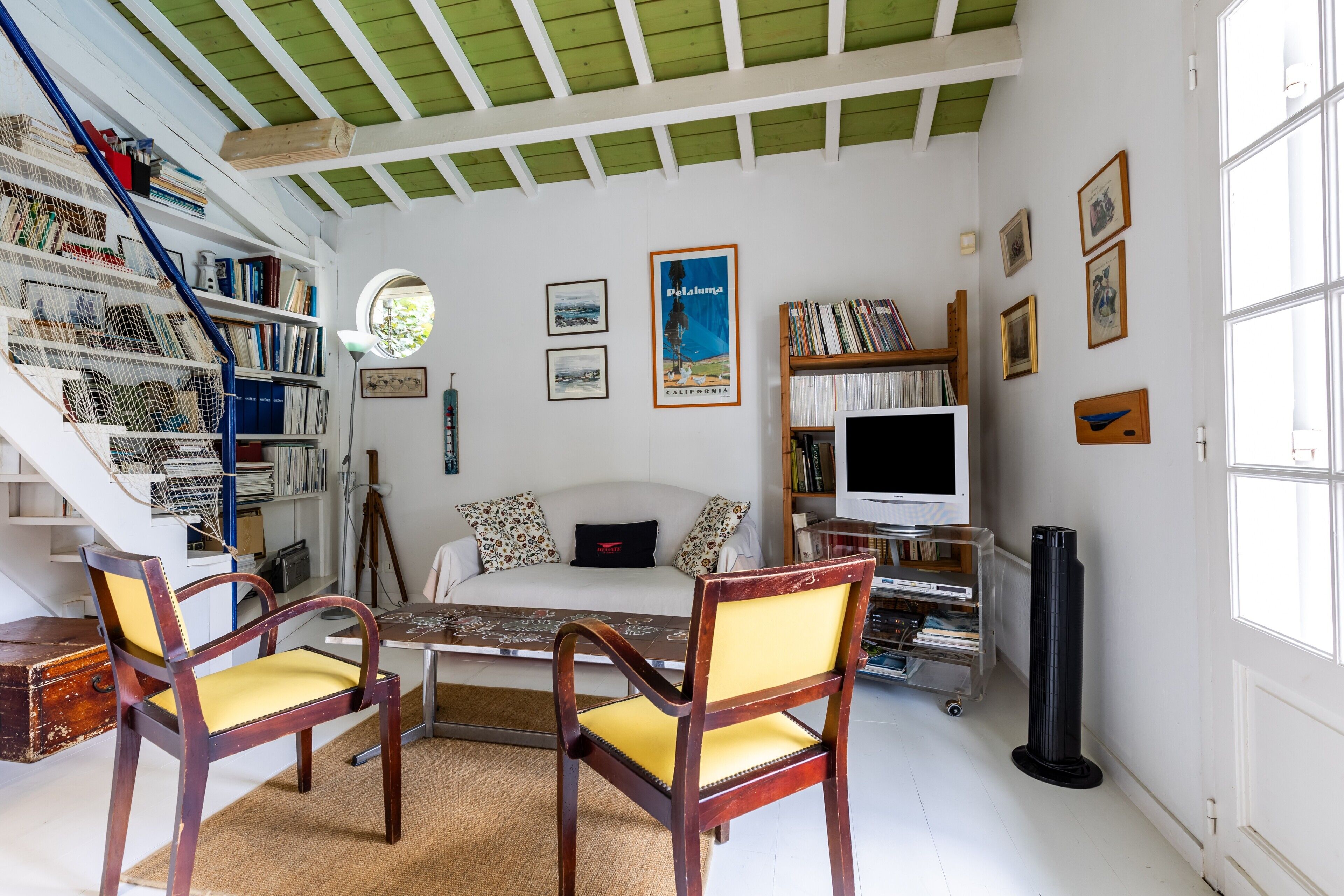 Ein Zimmer mit einem weißen Sofa, zwei Holzstühlen, einem Tisch, einem Bücherregal, einem Fernseher und einem Fenster.