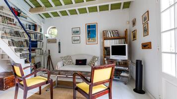 Ein Zimmer mit einem weißen Sofa, zwei Holzstühlen, einem Tisch, einem Bücherregal, einem Fernseher und einem Fenster.
