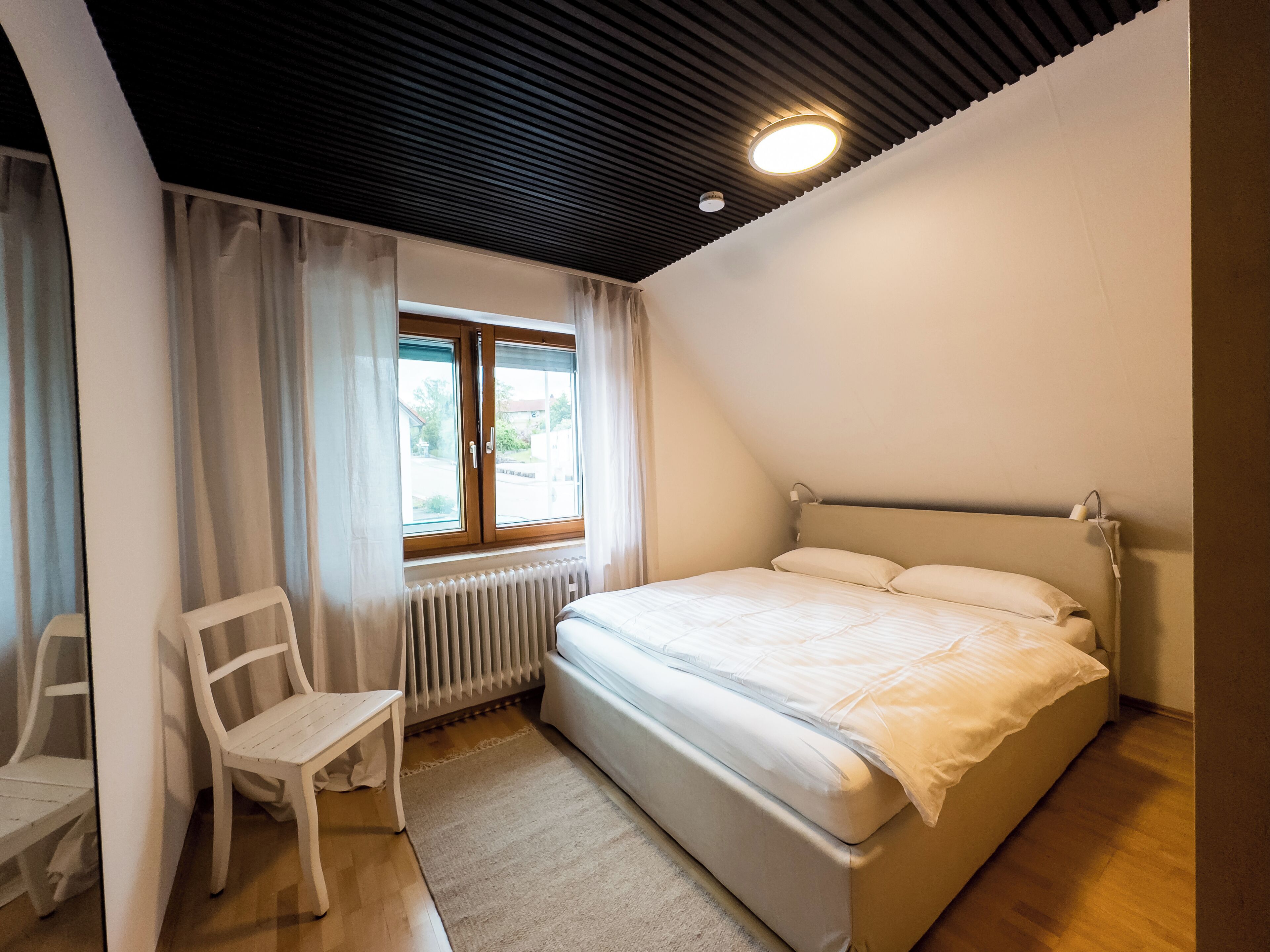 Un dormitori amb un llit, una cadira, una finestra amb cortines, un radiador i una làmpada al sostre.