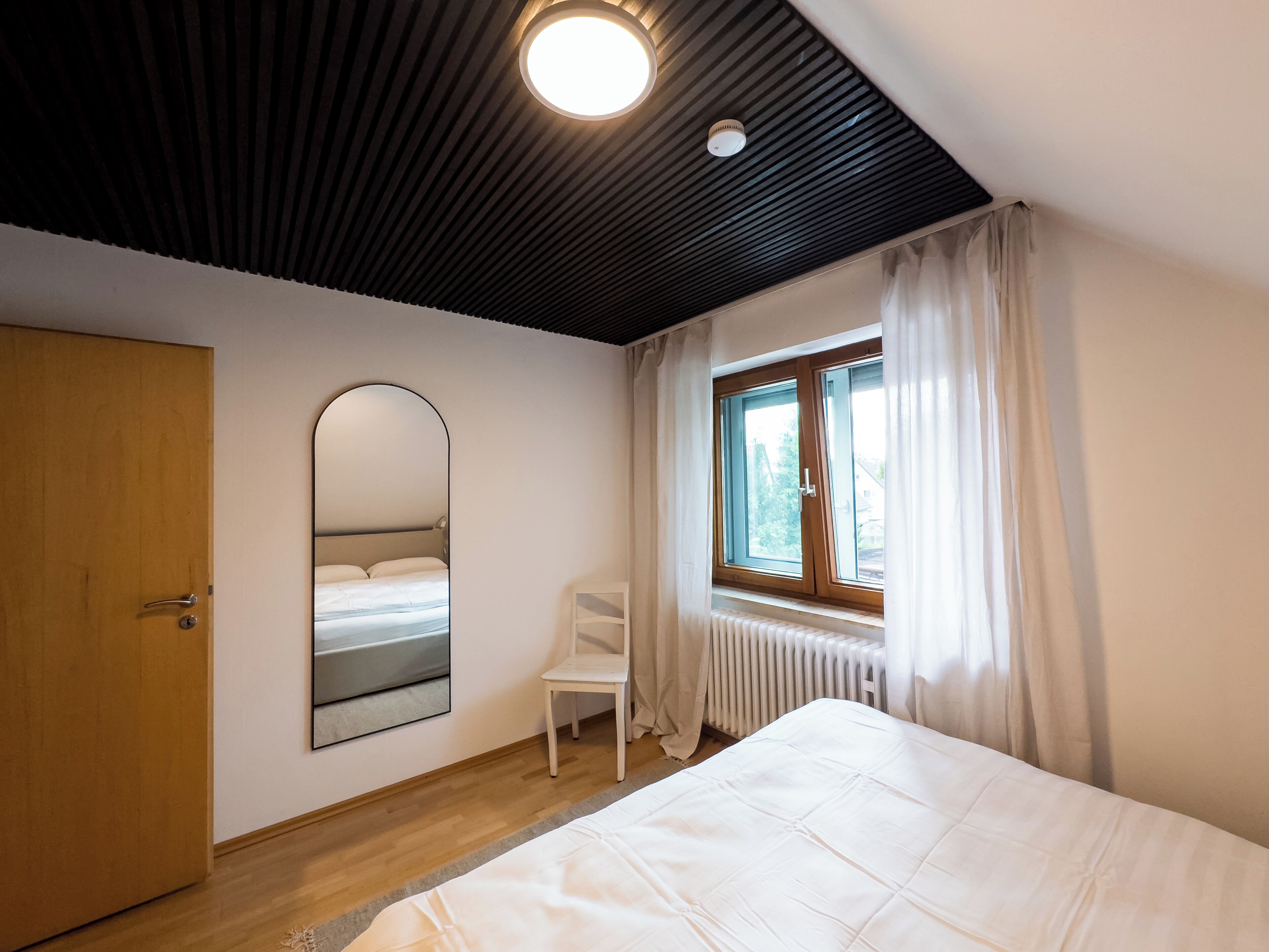 A bedroom with a bed, a chair, a window with curtains, a radiator, and a door.