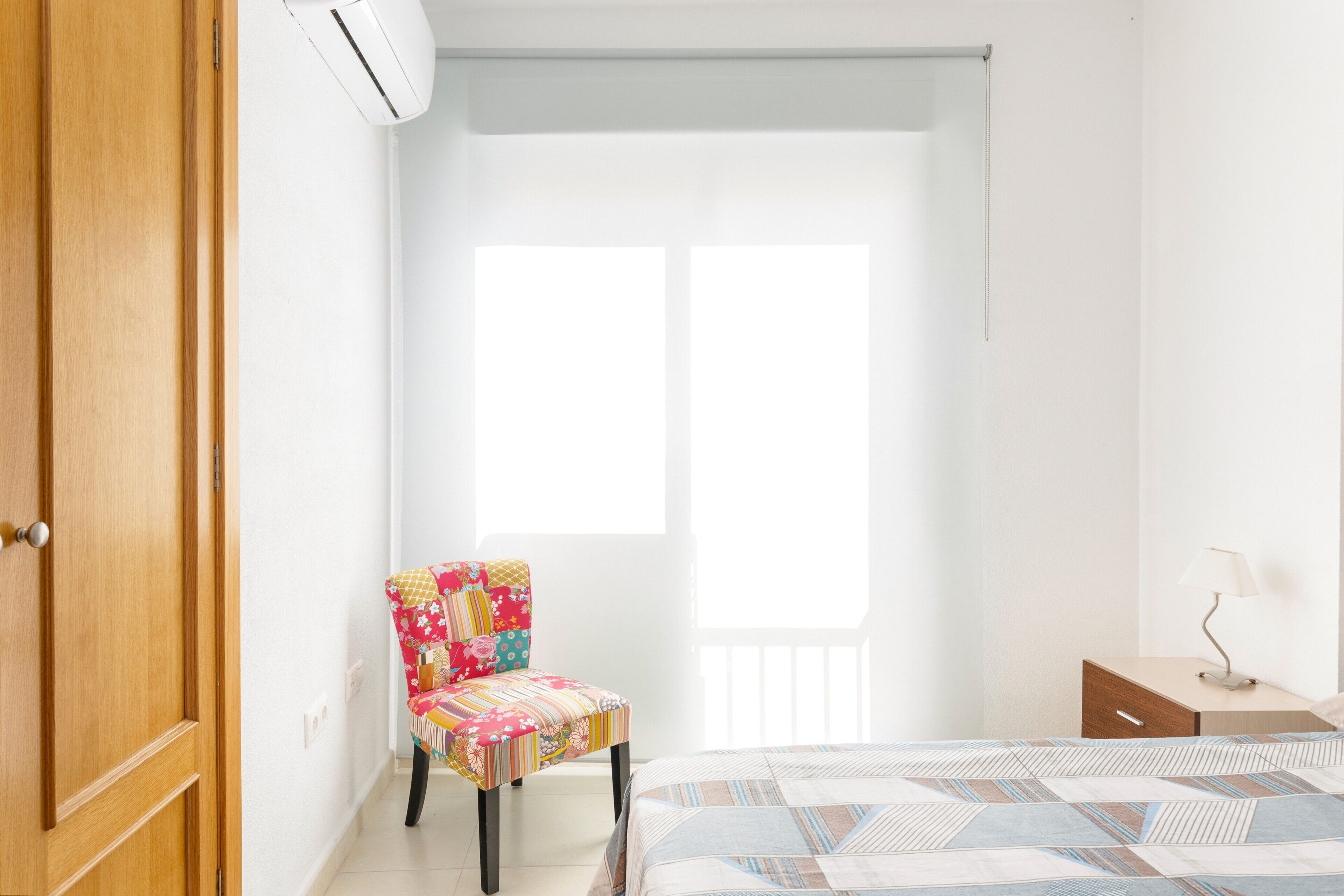 Un dormitorio con cama, una silla, una mesita de noche y una ventana.