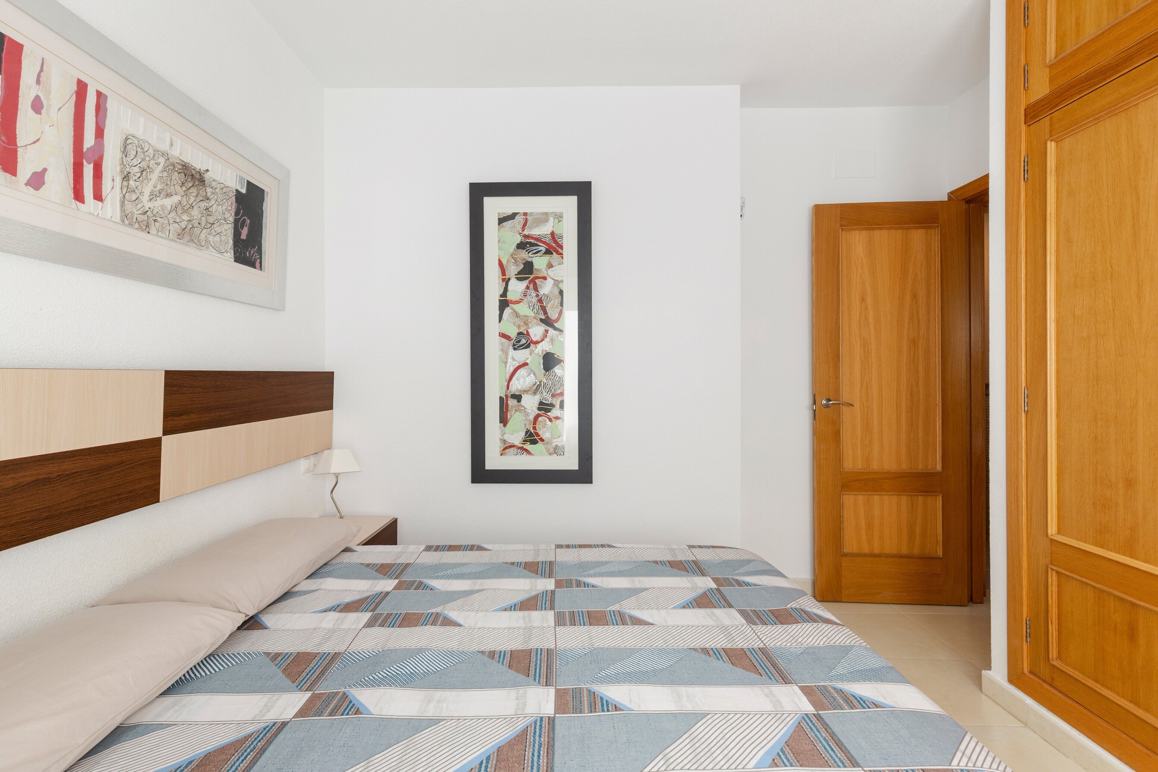 Un dormitorio con cama, un armario de madera, una puerta y una obra de arte abstracta enmarcada en la pared.
