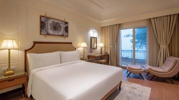 A hotel room with a large bed, two bedside lamps, a desk with a chair, a large window with curtains, and a decorative wall piece.