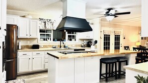 A modern kitchen with white cabinetry, a stainless steel refrigerator, a black range hood, and a ceiling fan. - 4BD | 3BTH | Pool | HotTub | Games |5Miles to I-95 (Elloree)