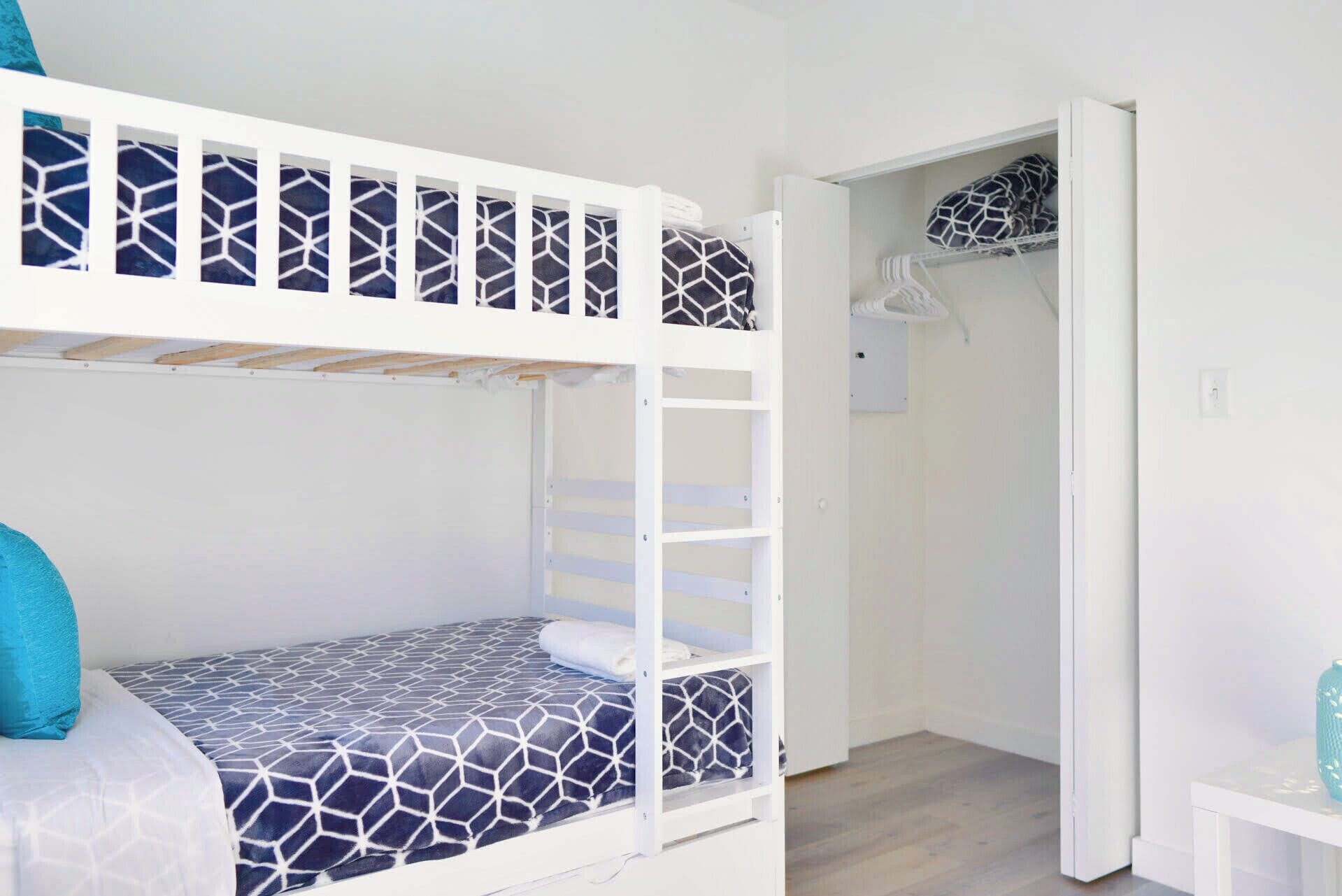 A bunk bed with a geometric-patterned mattress and pillows, a white wardrobe with a hanging organizer, and a small bedside table with a vase.