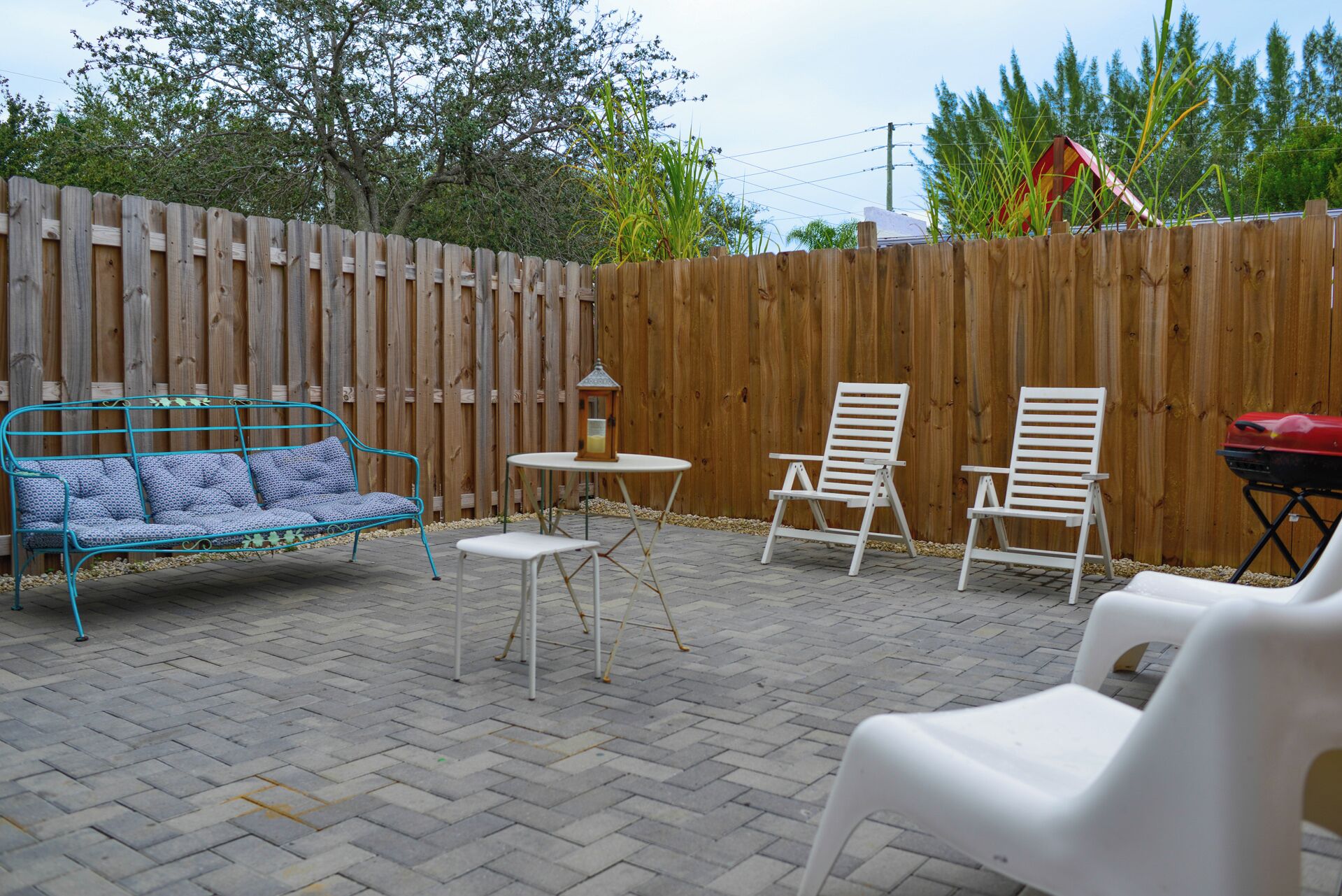 A patio with a wooden fence, a blue metal bench, two white folding chairs, a small round table, and a lantern.