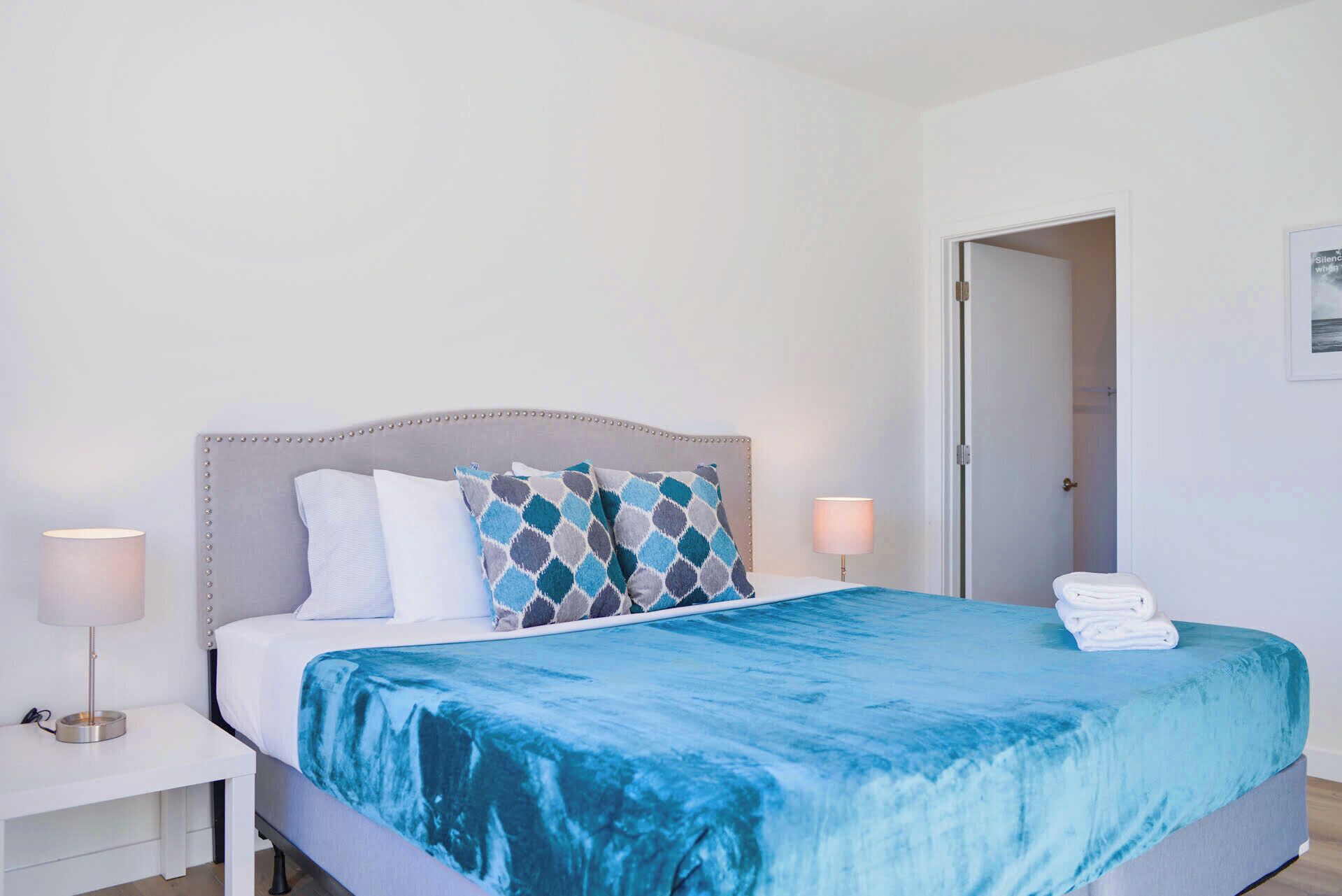 A neatly made bed with a blue blanket, two bedside lamps, and a door leading to another room.