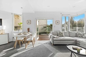 A modern living room with a sofa, a dining table with chairs, and a coffee table. There is a large window providing a view of the outdoors.