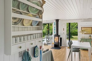 A modern interior with a white wall-mounted shelf holding plates and mugs, a black wood-burning stove, and a dining area with a table and chairs. - Holiday bliss near beach and nature (Græsted)