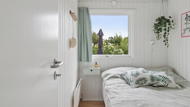 Et lille, enkelt værelse med seng, vindue med grønne omgivelser, sengebord og radiator.