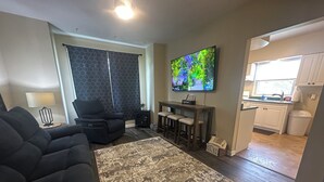 A living room with a black sofa, a patterned rug, a TV, and a bar area. - Spotless, Quiet 2BR Cottage: Private Deck, Big TV, Yard—Near Museums & Parks (New Brighton)