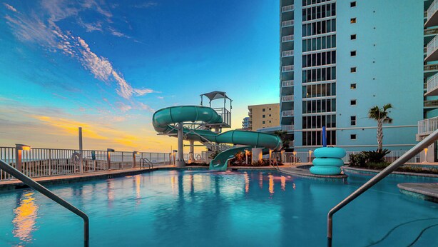 A swimming pool with a water slide, a tall building in the background, and a clear sky. - Phoenix Gulf Shores II 1002 (Gulf Shores)