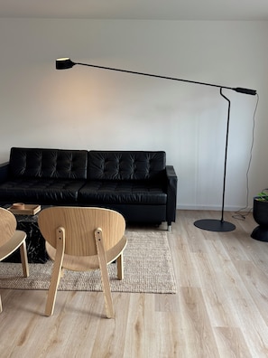 Uma sala de estar moderna com um sofá de couro preto, cadeiras de madeira, um abajur preto e uma mesinha com uma planta.