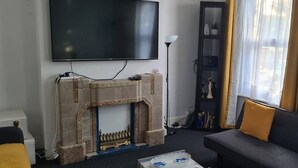 Una sala de estar con chimenea, un televisor de pantalla plana montado en la pared, un sofá con cojines y una lámpara de pie alta.