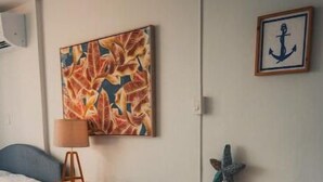 Une pièce comprenant un lit, un bureau et une table à manger avec des chaises. Il y a un tableau au mur et une photographie encadrée représentant une ancre.