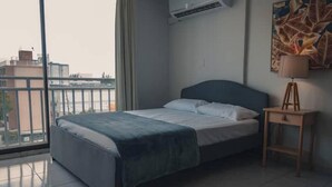 Une chambre d’hôtel moderne dotée d’une grande fenêtre, d’un lit avec une tête de lit bleue, d’une table de chevet avec une lampe et d’un climatiseur fixé au mur.