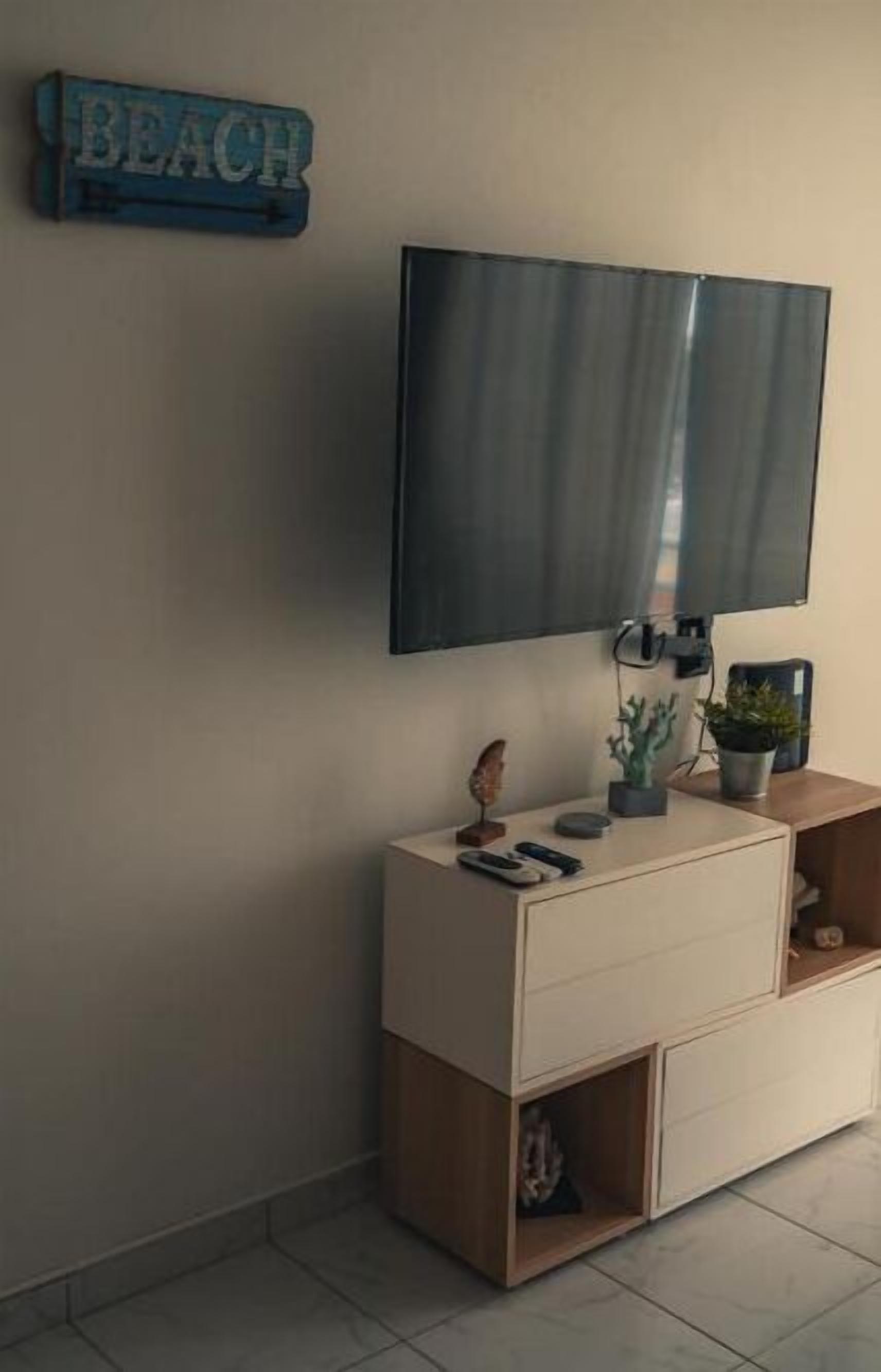 Un salon moderne avec un téléviseur à écran plat fixé au mur, un meuble en bois avec des tiroirs, une petite étagère ornée d’objets décoratifs et un panneau indiquant le mot « PLAGE ».