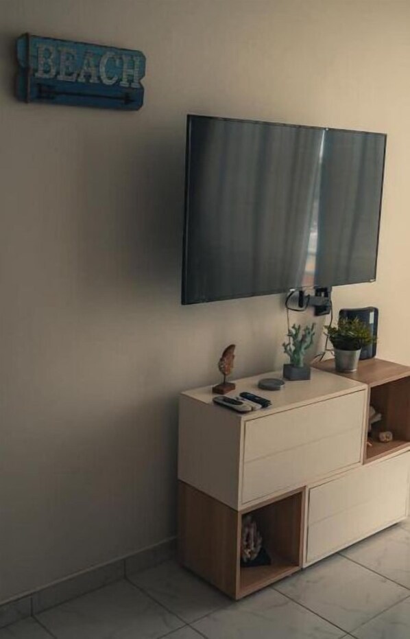 Een moderne woonkamer met een flatscreen televisie aan de muur, een houten kast met lades, een klein plankje met decoratieve spullen, en een bordje met daarop 'BEACH'.
