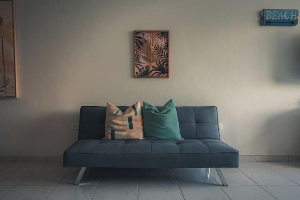 Een donkergrijze bank met twee kussens tegen een effen muur, met erboven een lijstwerk met een tropisch blad, en een bordje met de tekst 'BEACH' op de muur.
