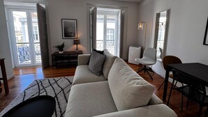 A modern living room with a sofa, a dining table, and a chair. There is a lamp on a side table, and a potted plant.