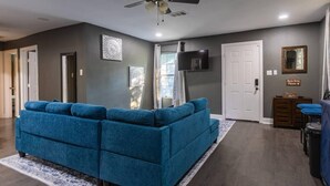 Een woonkamer met een blauwe modulaire bank, een plafondventilator, een flatscreen-tv en een eethoek met een bar.