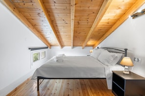 Een slaapkamer met een houten plafond, een bed met wit beddengoed, een nachtkastje met een lamp en een raam met uitzicht.