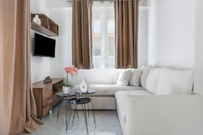 Een moderne woonkamer met een witte bank, een houten dressoir, een flatscreen-televisie, een kleine ronde tafel met twee stoelen, en een raam met gordijnen.