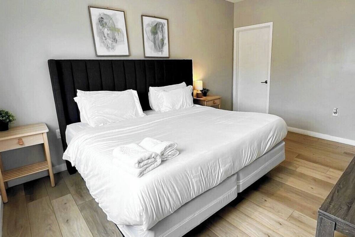Un lit bien fait, avec du linge de lit blanc et des oreillers, une table de chevet avec une lampe, et une porte sur la droite.
