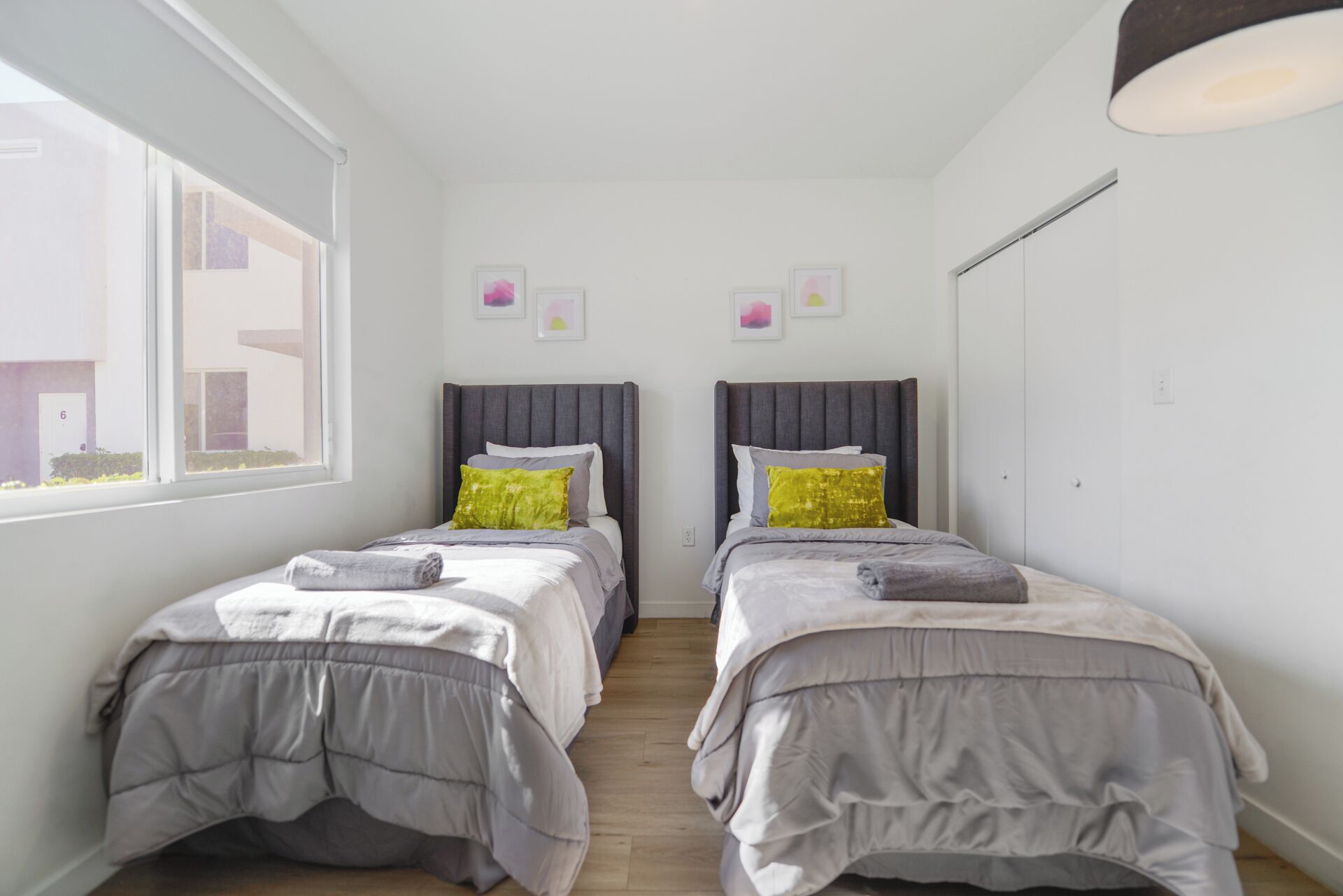 A bedroom with two beds, each with a grey headboard and a grey duvet. There are two pillows on each bed, one green and one white. The room has white walls and a window on the left side.