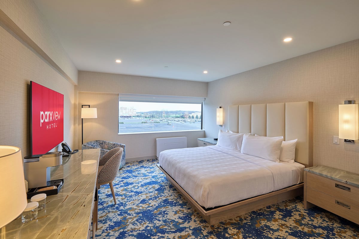 A hotel room with a bed, bedside tables, a desk with a chair, a large window, and a view of the city.