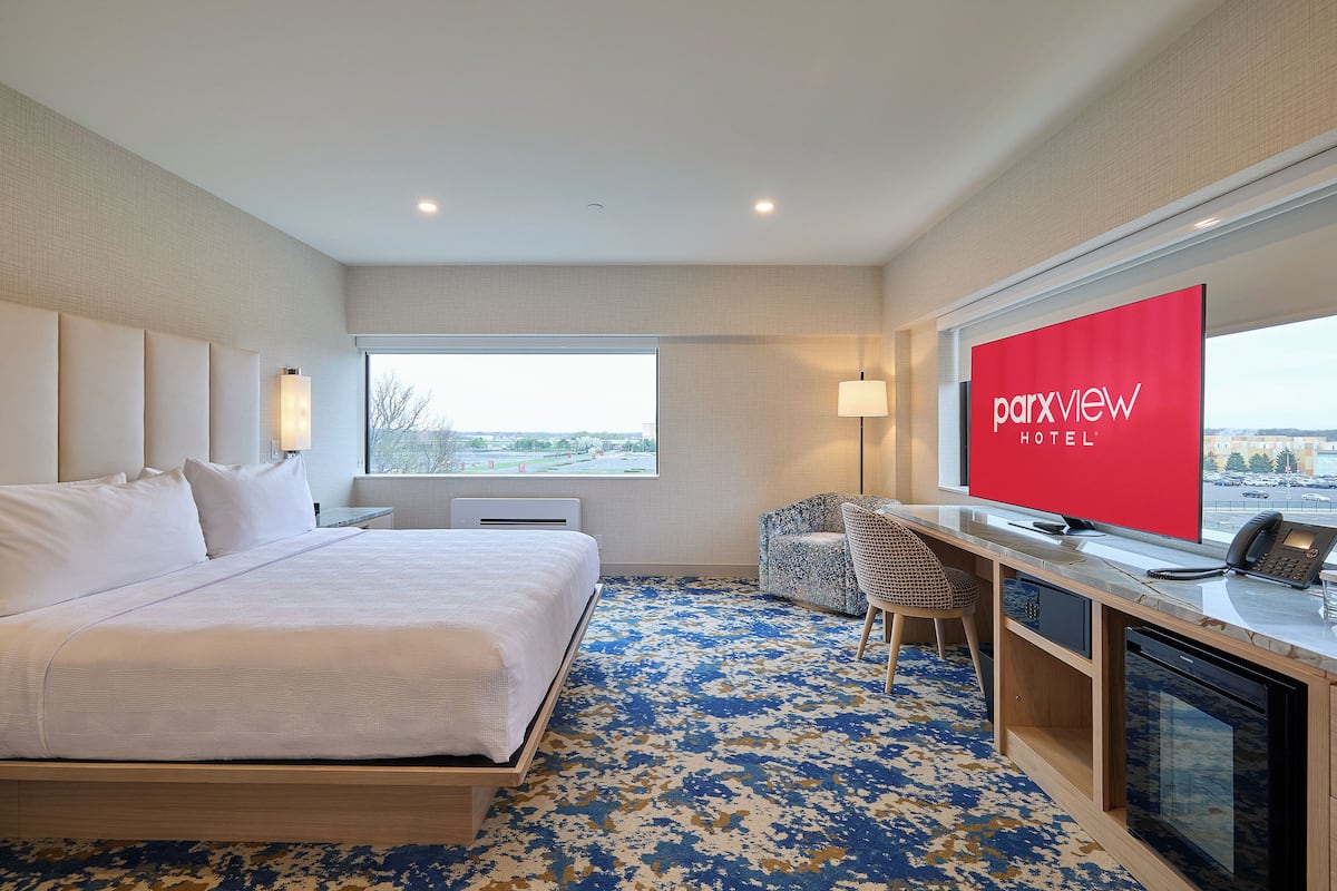 A hotel room with a bed, desk, chair, TV, and a view of the city.