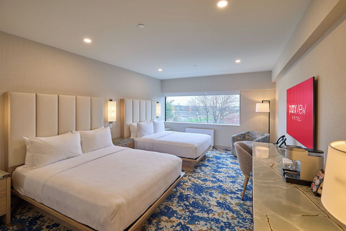 A hotel room with two beds, a desk with a lamp, a chair, and a large window.