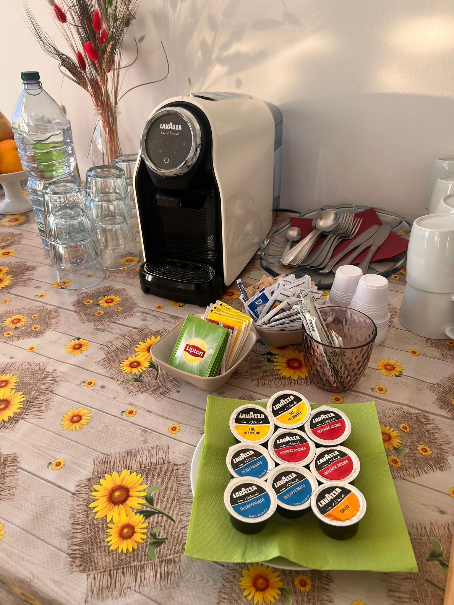 Una mesa con una cafetera, vasos, cubiertos y diferentes cápsulas de café.