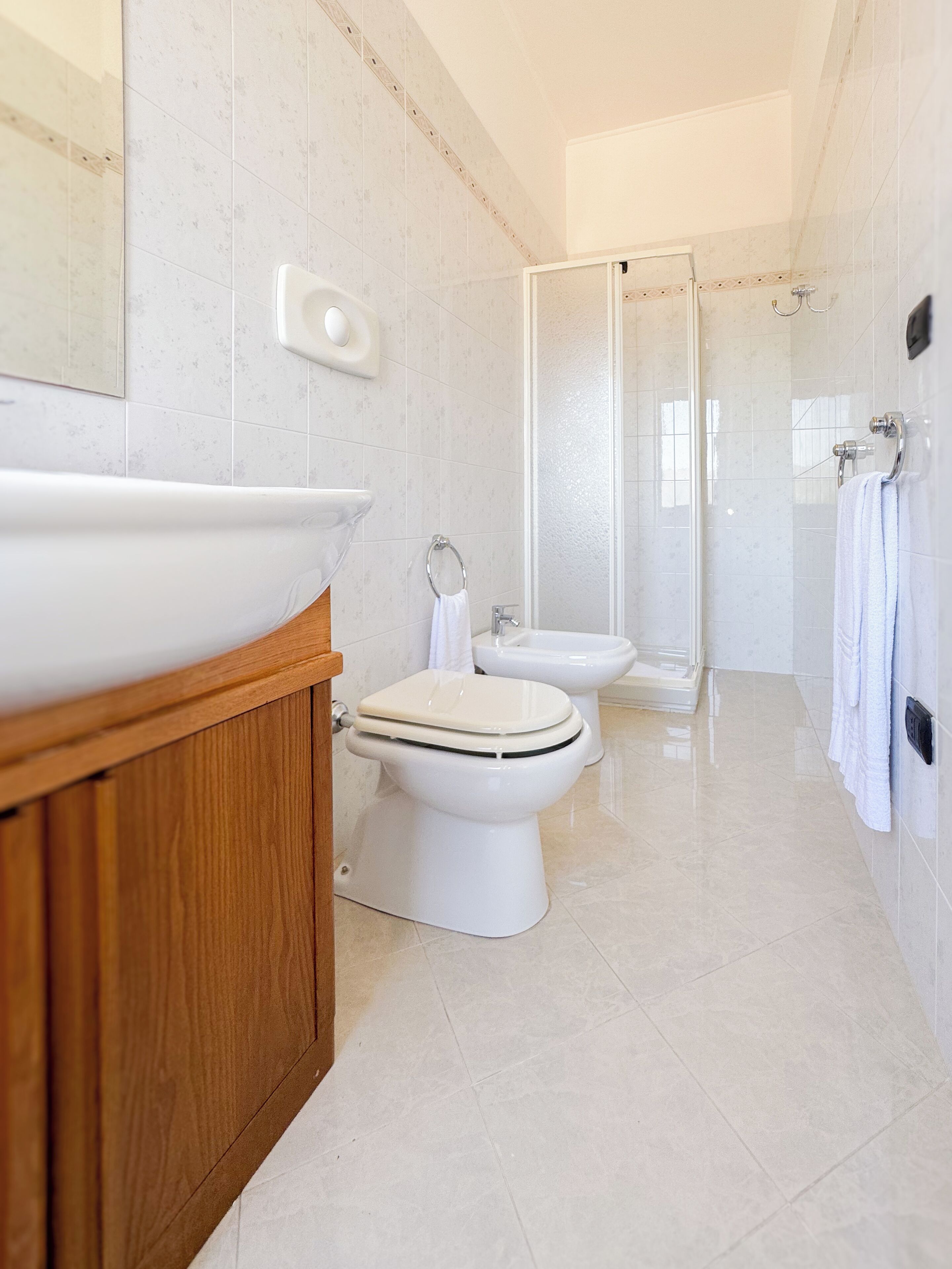 Ein modernes Badezimmer mit einem großen weißen Waschbecken, zwei Toiletten, einer Duschkabine aus Glas und einem hölzernen Waschtisch.