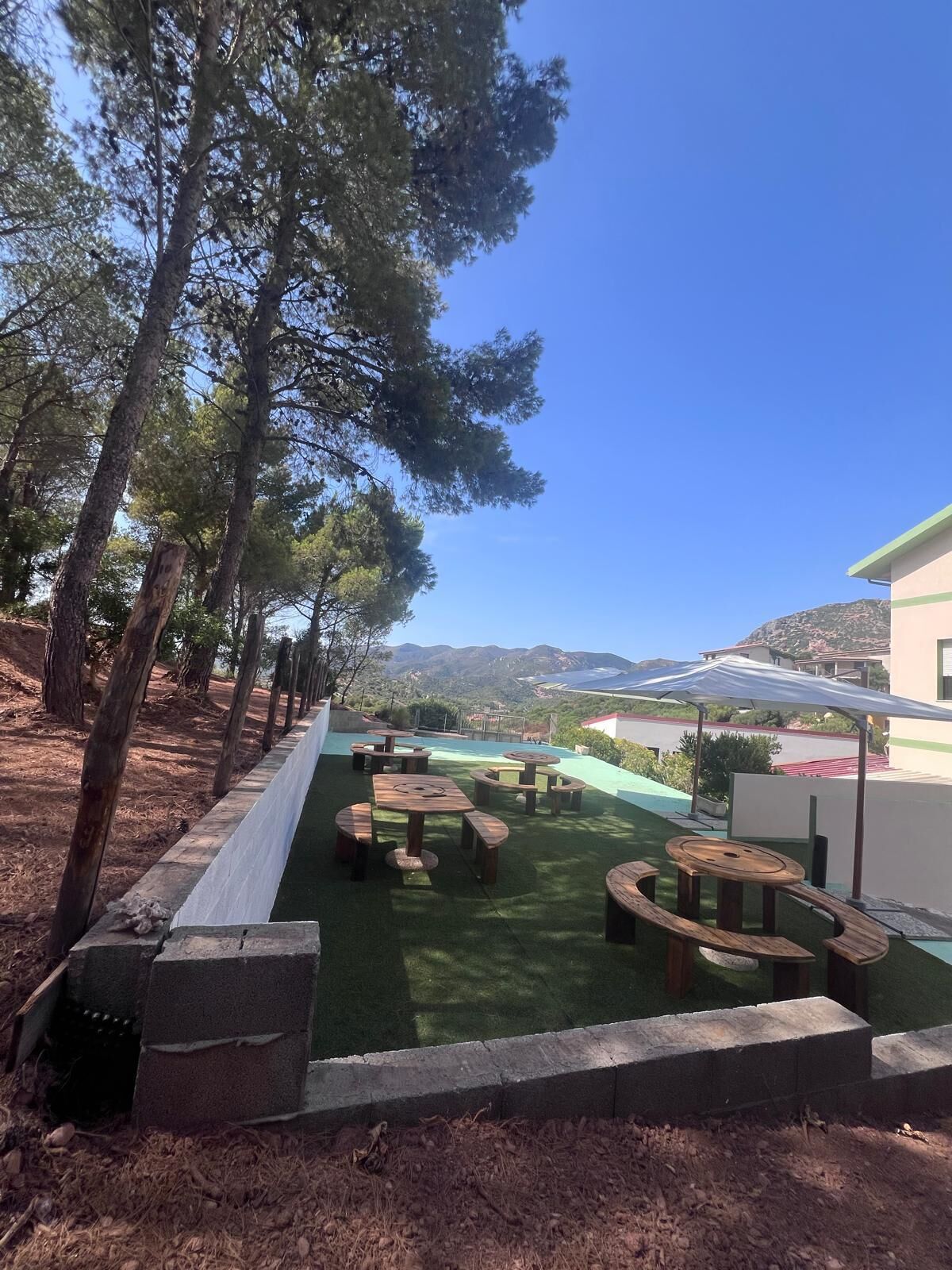 A patio area with wooden tables and chairs, surrounded by trees and a view of the landscape.