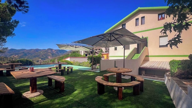 An outdoor patio with wooden tables and benches, a pool, and a building with a green roof and a staircase. - Ostello Brebei Niedda (Gonnesa)